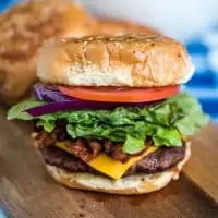 bacon cheeseburger with lettuce, onions, and tomatoes on wooden cutting board on picnic table