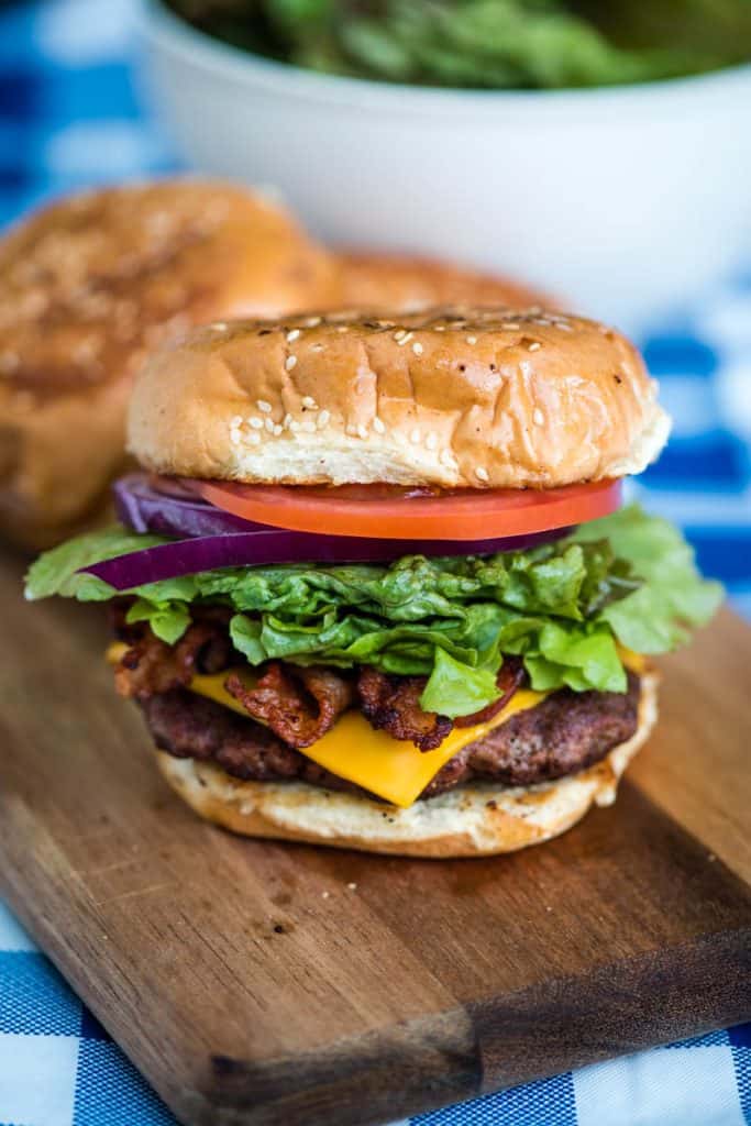 bacon cheeseburger with lettuce, onions, and tomatoes on wooden cutting board on picnic table