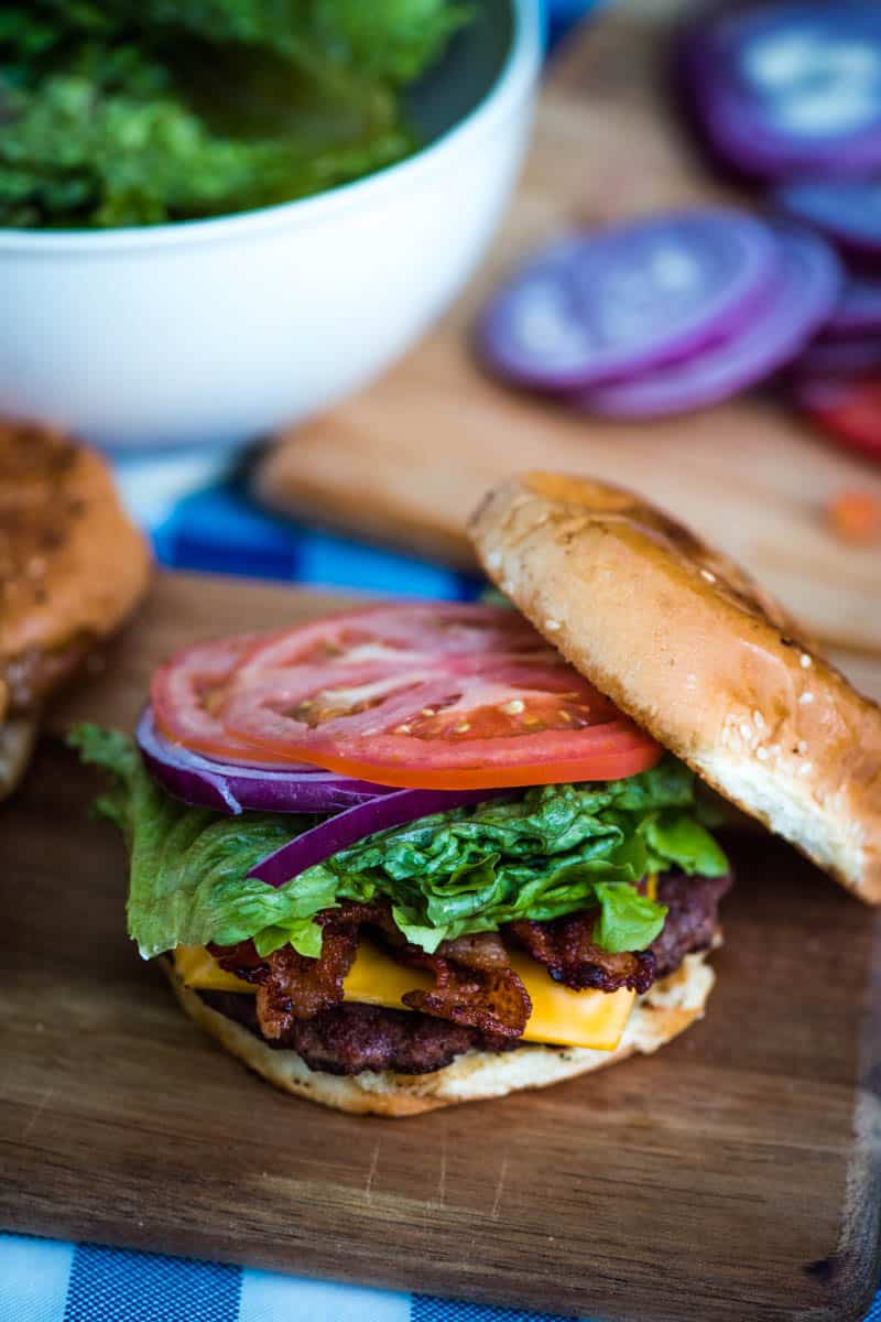 ultimate bacon cheeseburger on wood cutting board with toppings, including lettuce, red onion, and tomato slices