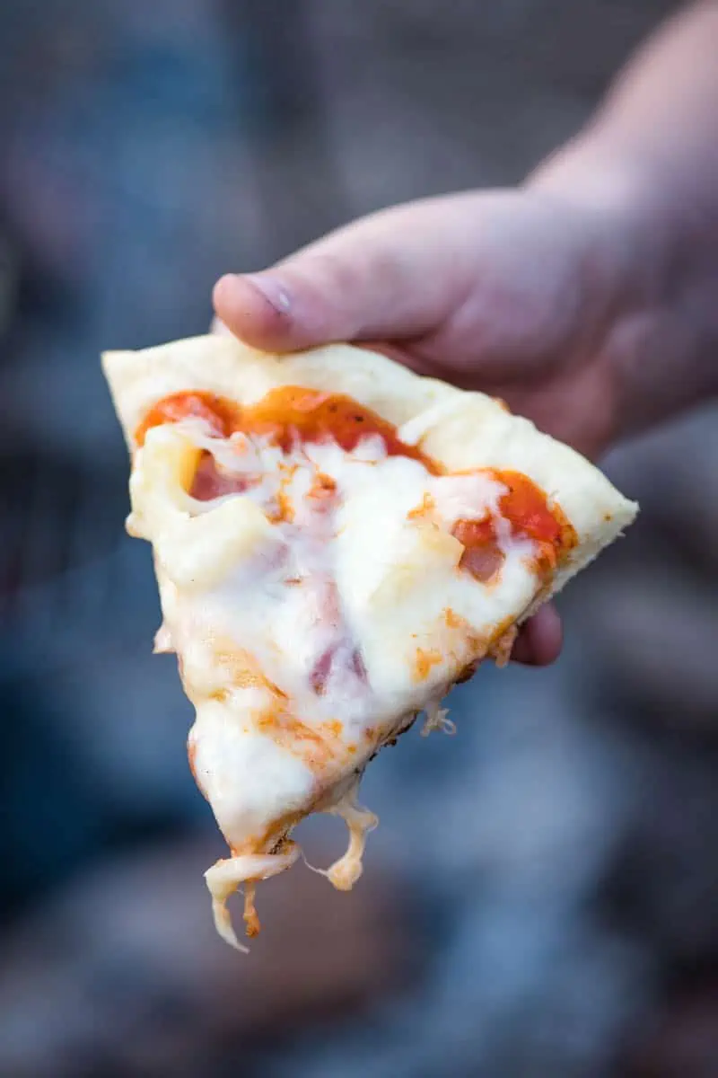 hand holding slice of Hawaiian pizza cooked over a campfire