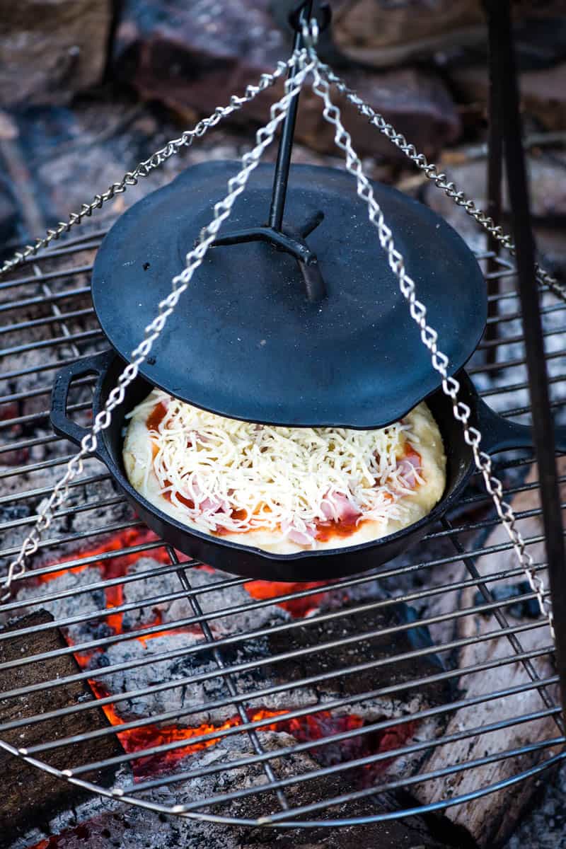 cooking ham and pineapple pizza on a campfire in a cast iron skillet with the lid