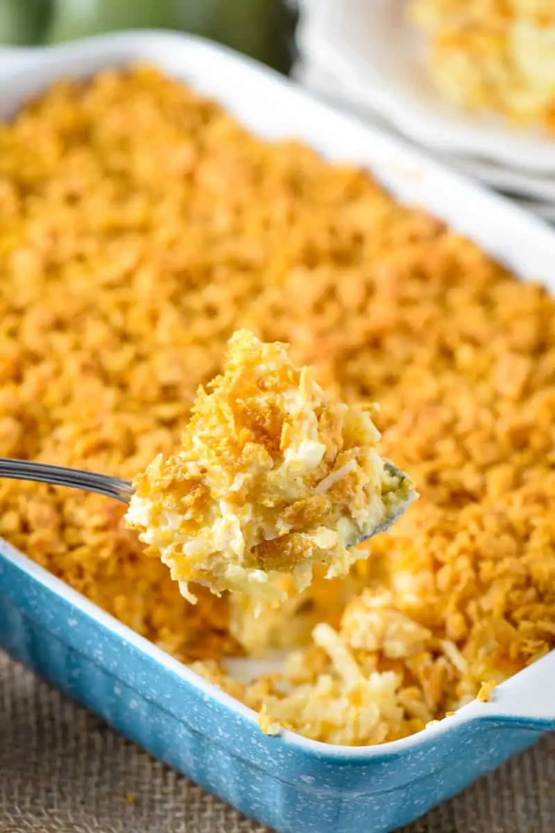 large spoonful of cheesy hash brown casserole with Corn Flakes above blue baking dish