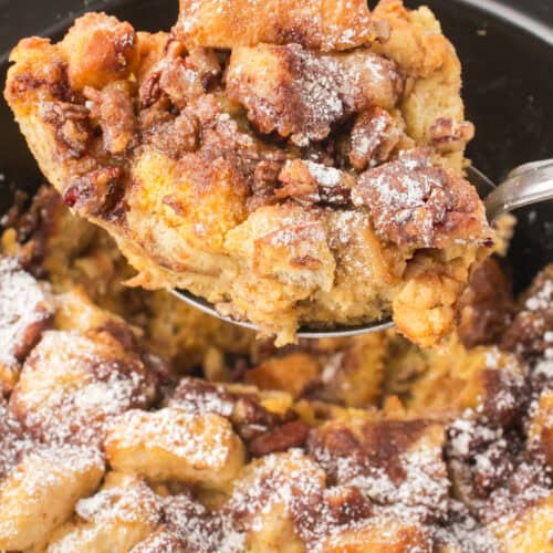 holding up a slice of French toast casserole over a black slow cooker full of French toast casserole