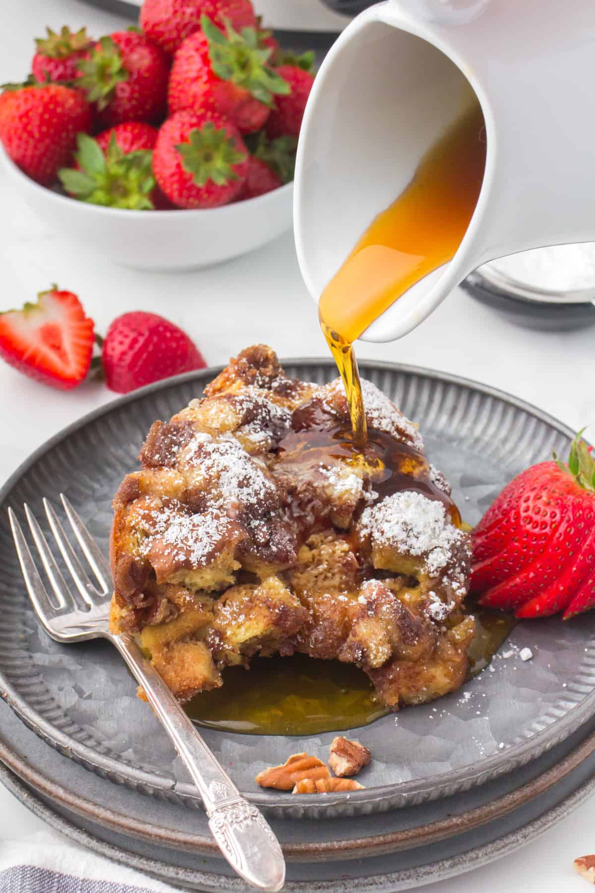 pouring maple syrup over a slice of slow cooker French toast casserole on a decorative gray plate with fresh strawberries on the side