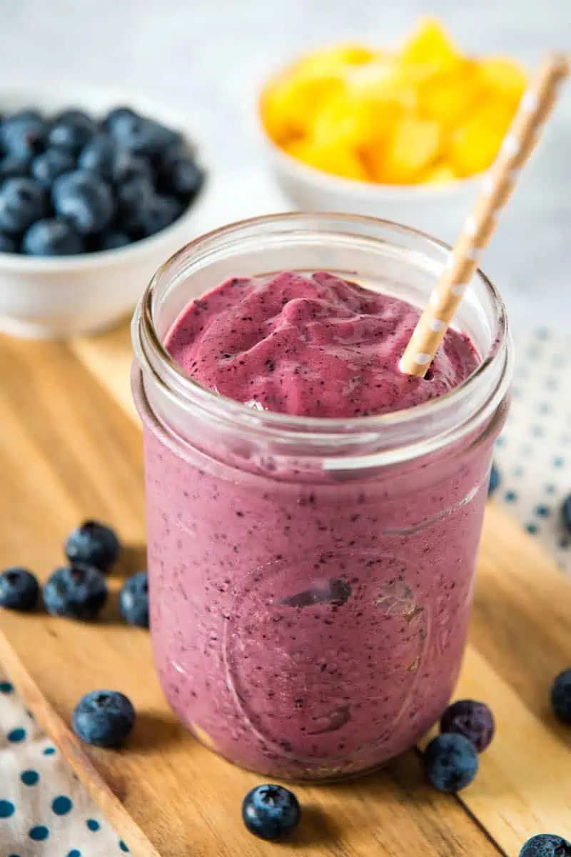 freshly made blueberry mango smoothie in wide mouth mason jar with brown straw, on wooden cutting board, with blueberries and mango in bowls behind