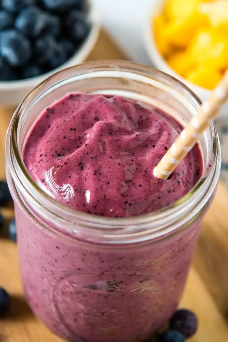 mango banana blueberry smoothie in wide mouth mason jar with brown paper straw, sitting on wood cutting board with blueberries