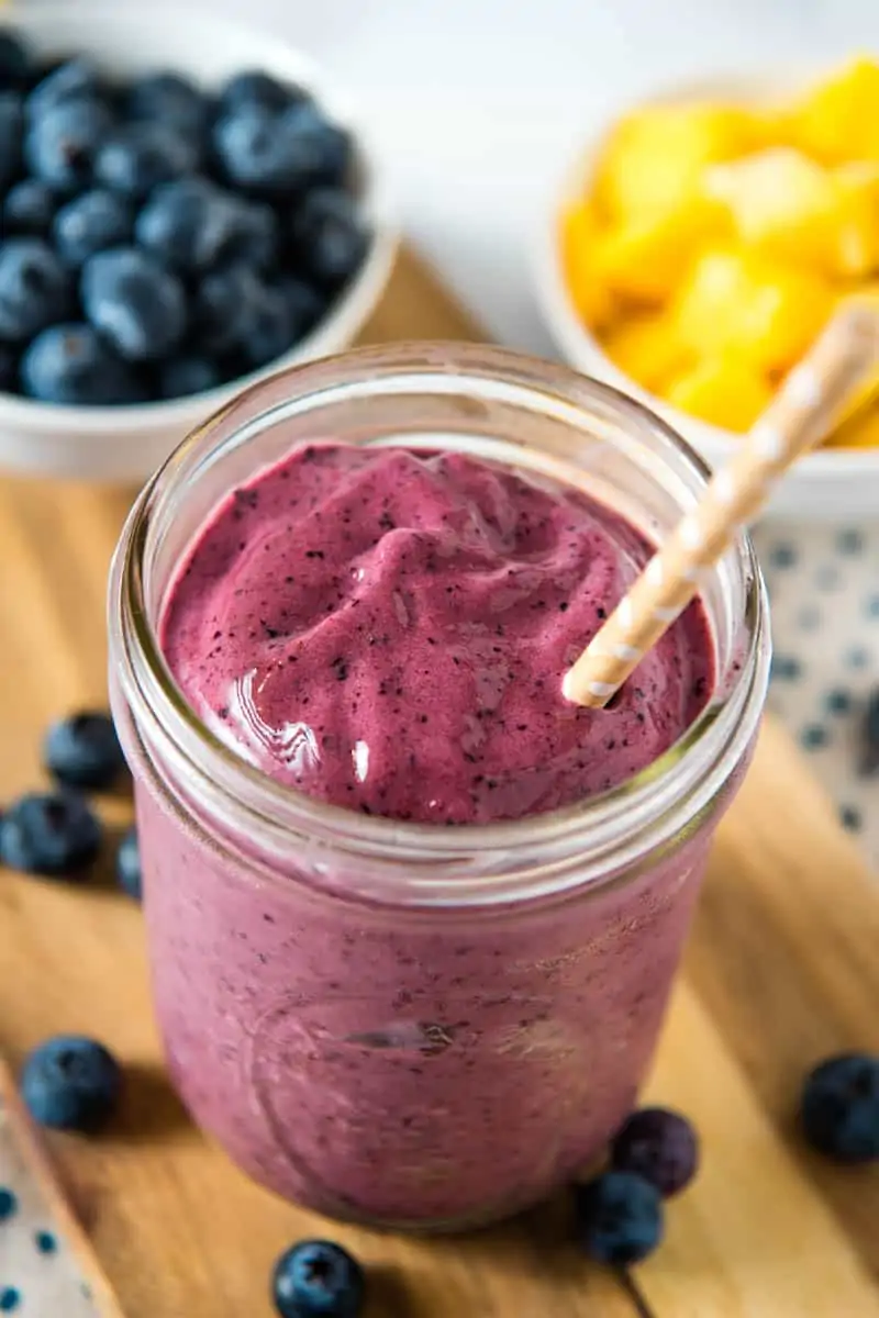 mango blueberry smoothie in glass Mason jar with brown paper white polka dot straw, on a wooden cutting board, with fresh blueberries and frozen mango in bowls