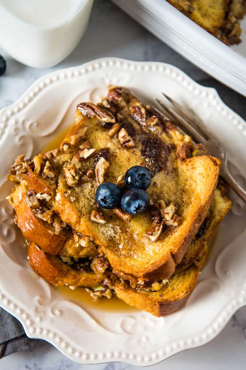 stack of baked brioche French toast casserole topped with blueberries, pecans, and maple syrup on white plate with fork