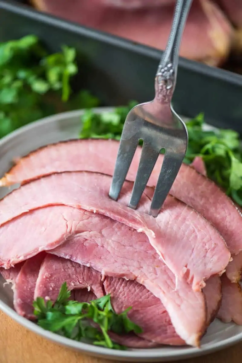 slices of baked ham on gray plate with serving fork stuck in ham