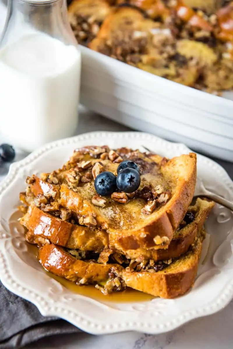 brioche French toast casserole with blueberries stacked on white plate with fork, gray napkin, and milk in jar