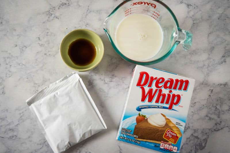whipped dessert topping ingredients, including Dream Whip whipped topping mix, milk, and vanilla extract on white marble countertop