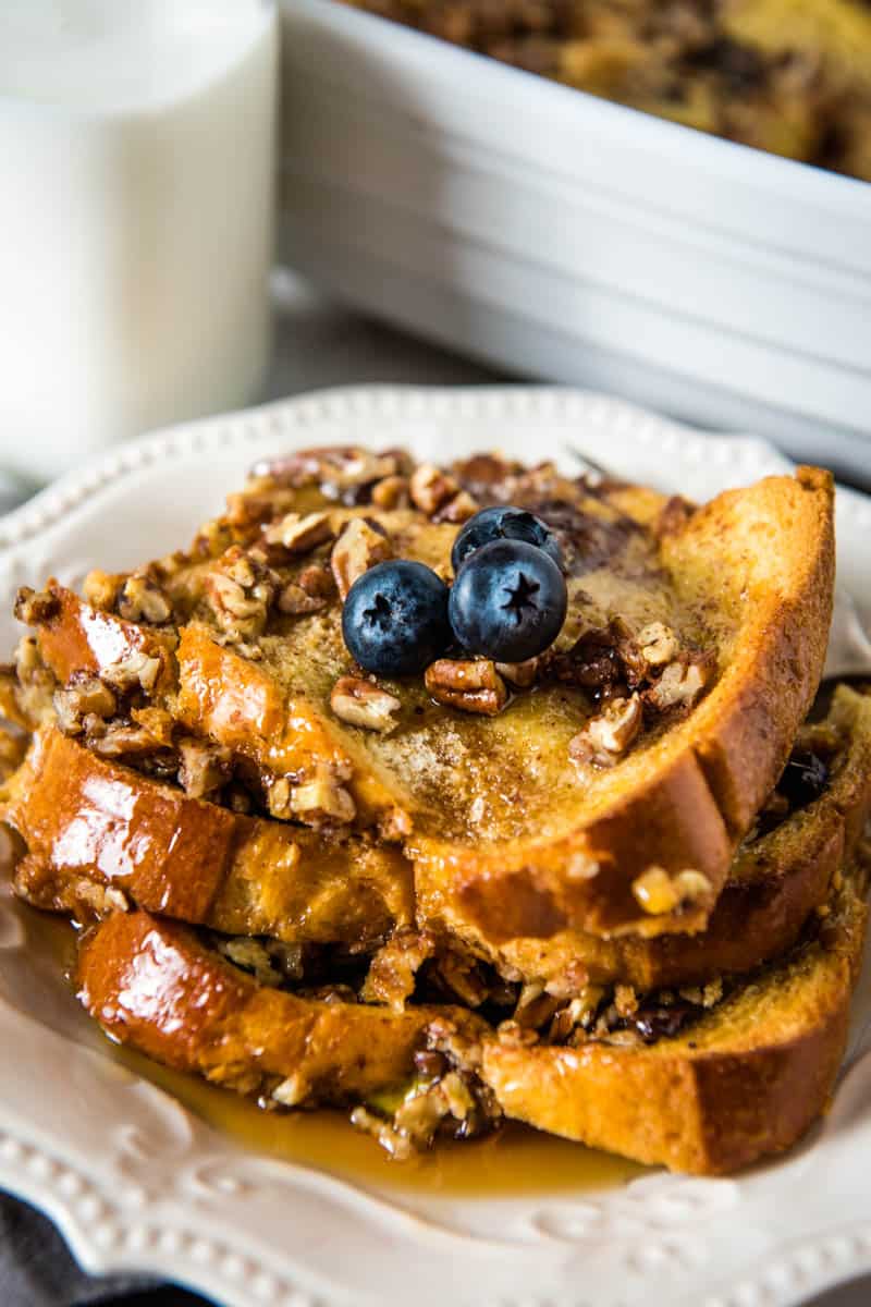 close up on stack of brioche French toast casserole, with pecans and blueberries, on white plate with fork and jar of white milk behind