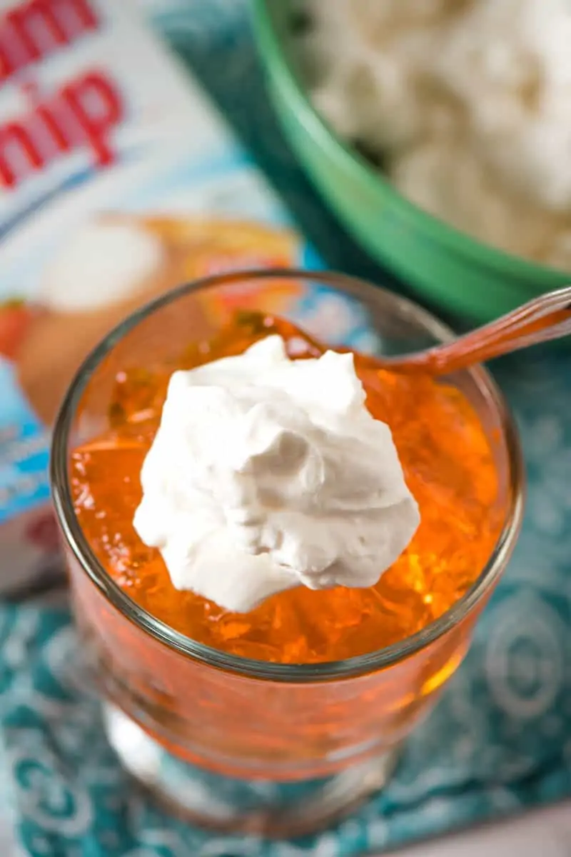 Orange Jello topped with Dream Whip whipped topping in glass dish with spoon on teal blue napkin with Dream Whip box