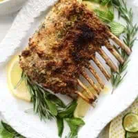herb crusted rack of lamb on rectangular white platter with fresh lemon and herbs