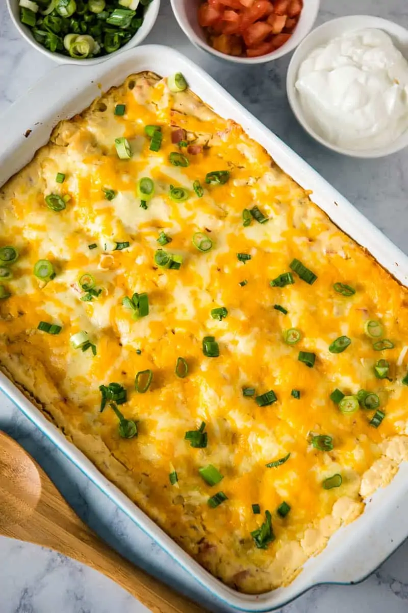 baked white sauce crab enchiladas in blue and white baking dish, surrounded by sour cream, tomatoes, green onions, and a wooden spoon