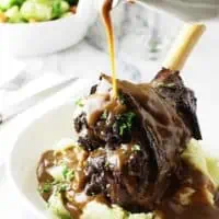 pouring brown gravy over braised herb lamb shanks in white bowl