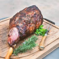 grilled leg of lamb on wooden cutting board with knives and fresh herbs