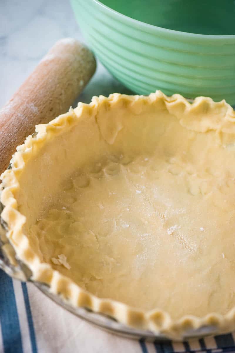 Butter pie crust in glass pie plate on blue striped white kitchen towel with wooden rolling pin and green mixing bowl