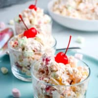 ambrosia fruit salad in small glass serving bowl on light blue plate with maraschino cherry on top