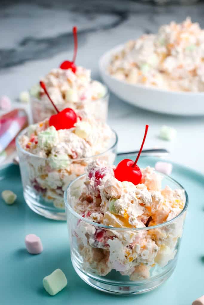 ambrosia fruit salad in small glass serving bowl on light blue plate with maraschino cherry on top