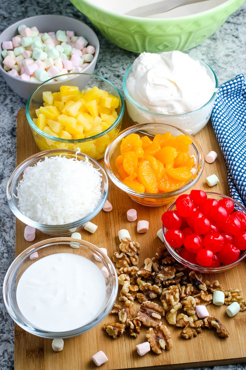 ambrosia ingredients, including cherries, whipped cream, mandarin oranges, pineapple tidbits, coconut, sour cream, multi-colored mini marshmallows, and nuts in small glass bowls on wooden cutting board
