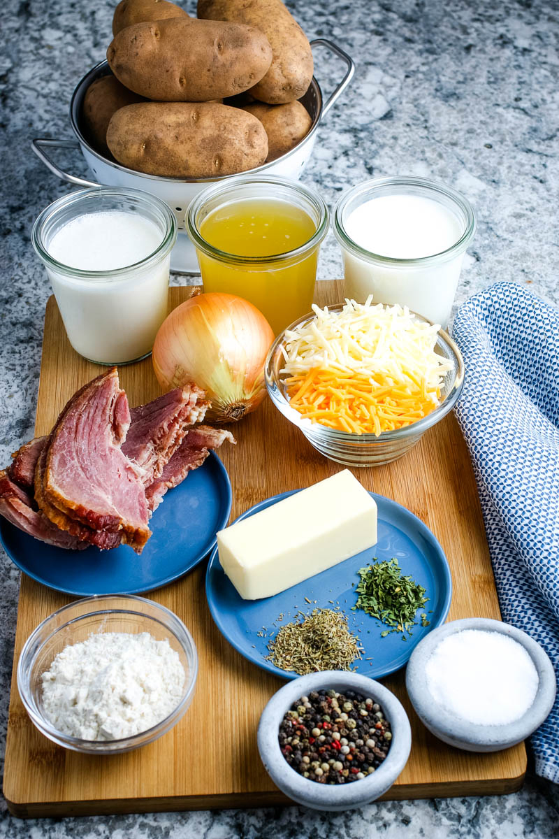 ingredients for scalloped potatoes and ham recipe, including potatoes, heavy cream, milk, chicken broth, yellow onion, cheddar cheese, Monterey Jack cheese, ham, butter, thyme, parsley, salt, pepper, and flour