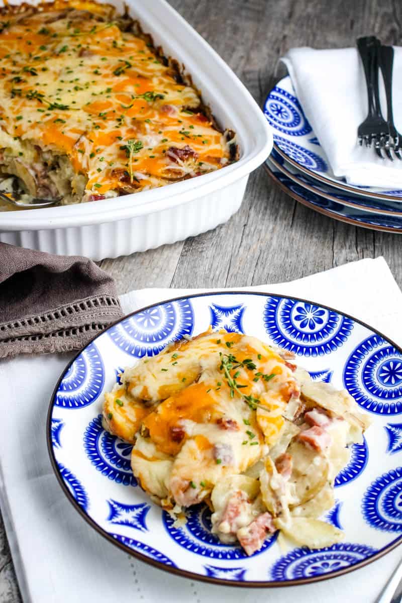 serving of scalloped potatoes and ham casserole on blue and white decorative plate with whole casserole in white casserole dish behind plate, along with more plates and forks for serving