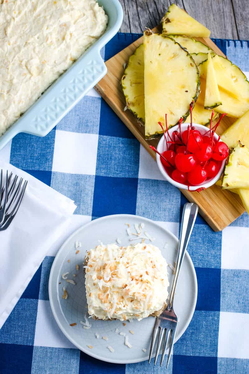 southern pineapple cake in pan with fresh pineapple and maraschino cherries on wood cutting board, and slice of pineapple cake, topped with frosting and shredded coconut, on light blue plate with fork