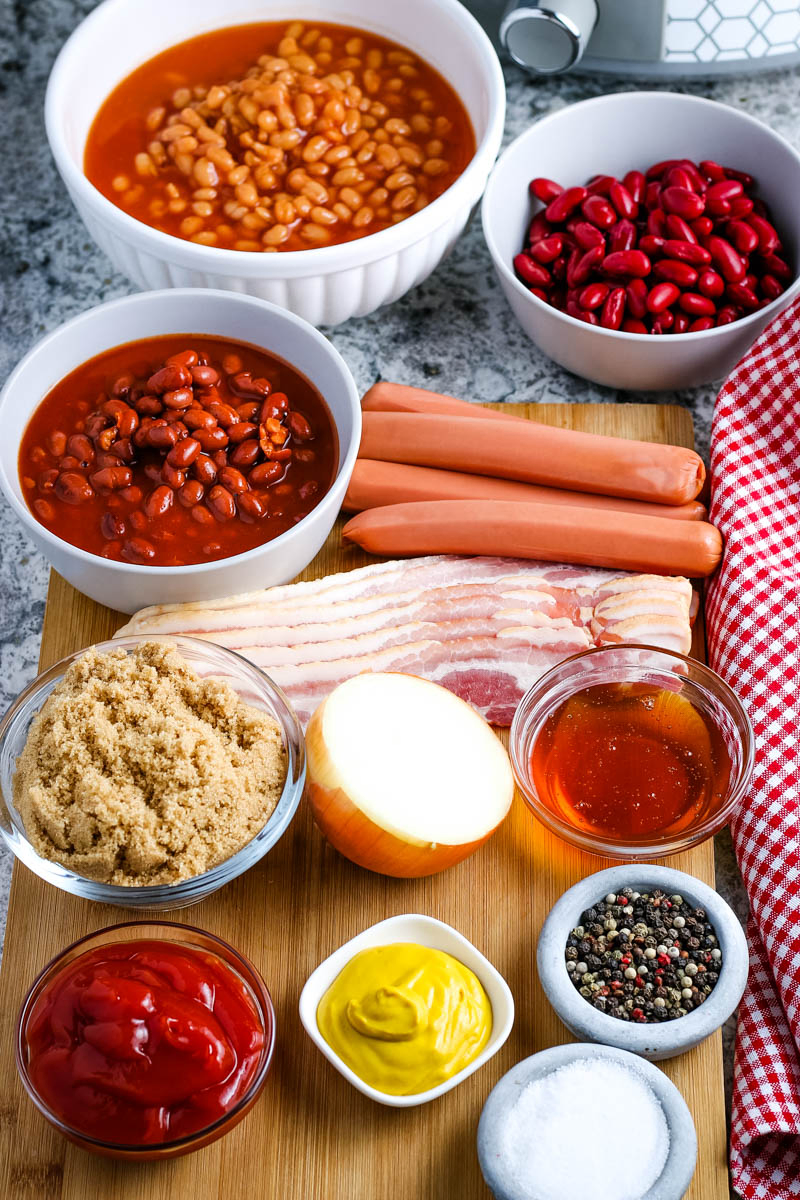 ingredients for baked beans in various bowls on wooden cutting board, including pork and beans, red kidney beans, chili beans, hot dogs, bacon, brown sugar, yellow onion, honey, ketchup, yellow mustard, salt, and pepper