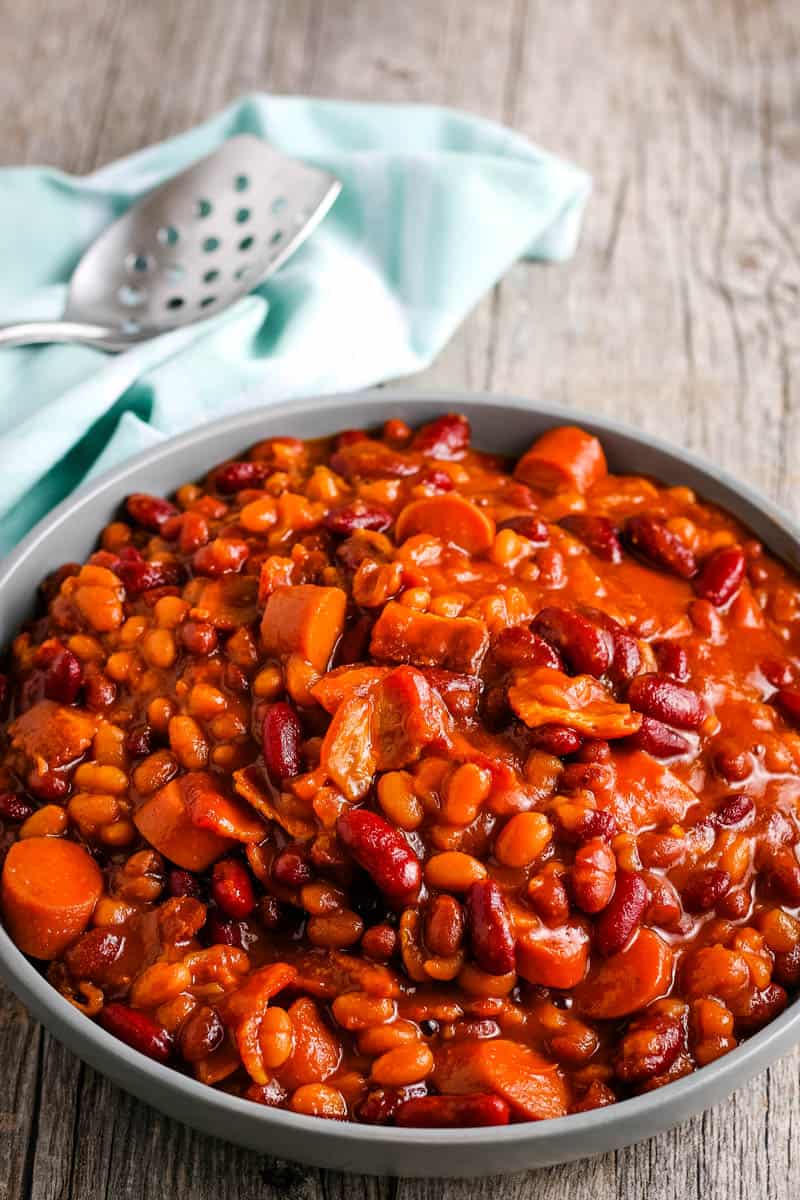 gray bowl full of homemade baked beans on wooden countertop with light teal kitchen towel and large holed serving spoon
