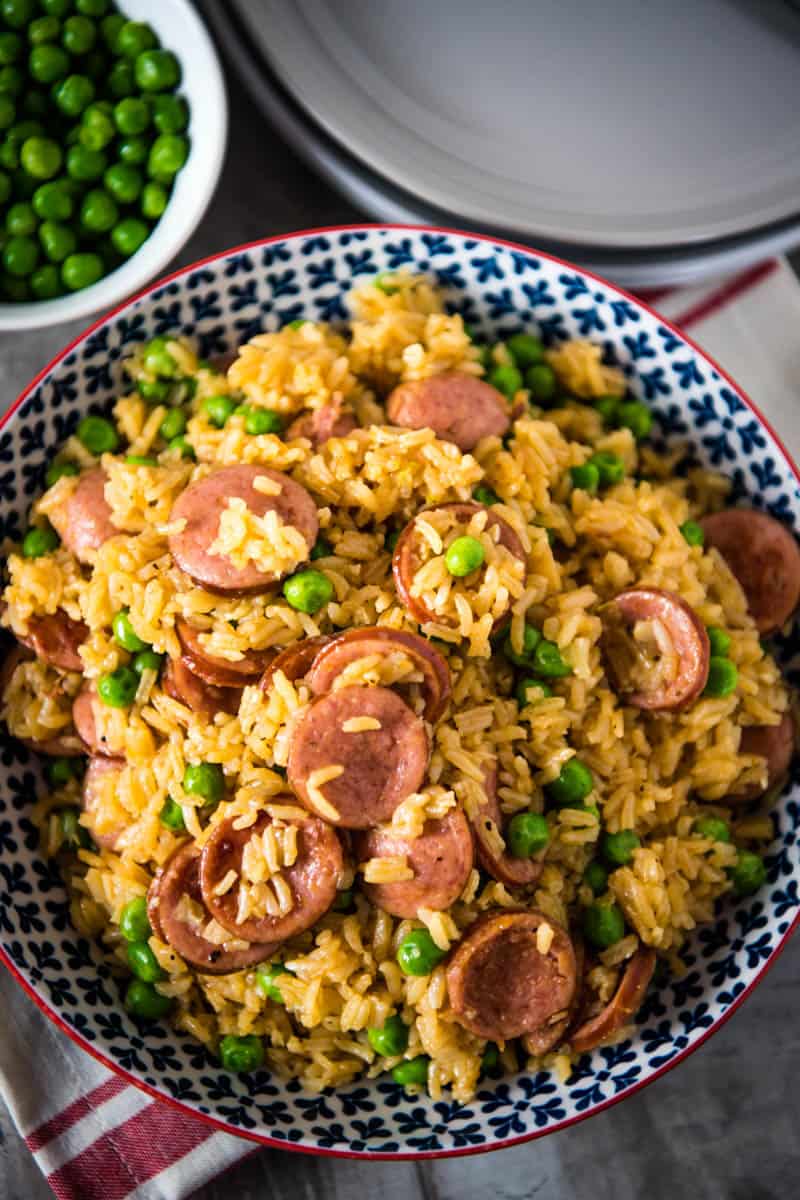 blue bowl full of pressure cooker sausage and rice with green peas, on wooden countertop with red and white striped kitchen towel