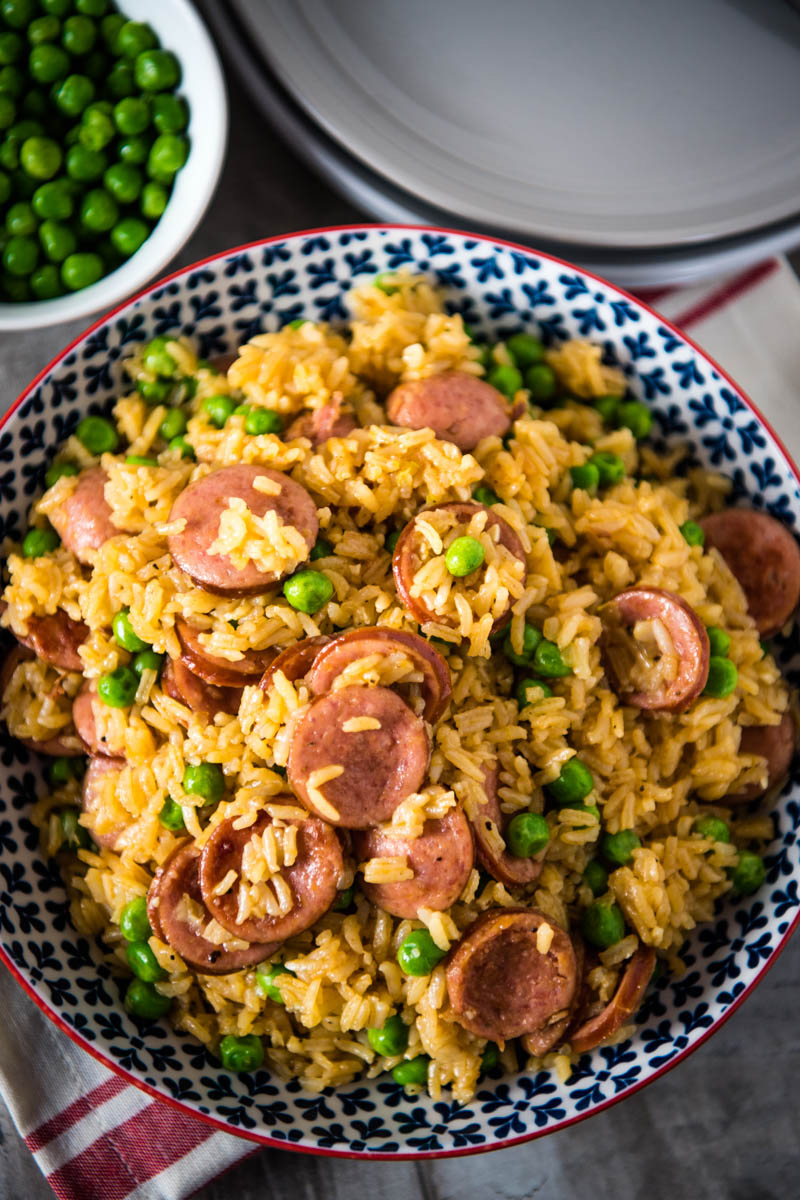 blue bowl full of pressure cooker sausage and rice with green peas, on wooden countertop with red and white striped kitchen towel
