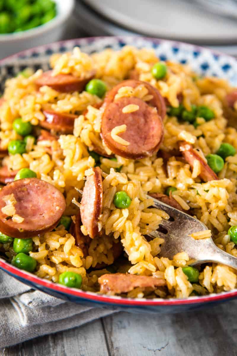 forkful of smoked sausage and rice, with green peas, in blue and red decorative bowl on wooden countertop with gray linen