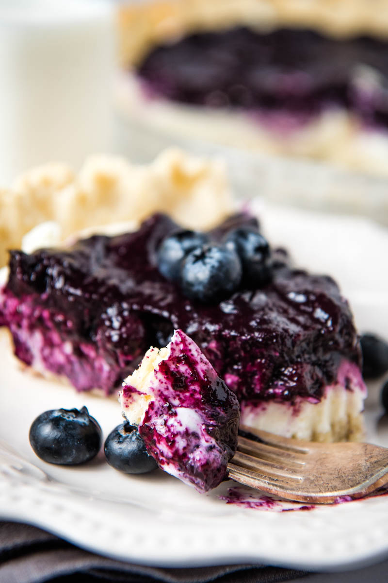 bite of blueberry cream pie on fork, with slice and fresh blueberries on white plate