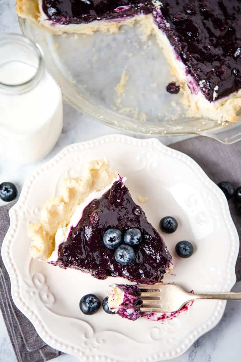 blueberry pie sliced and served on white plate with fresh blueberries and a fork, along with a jar of milk