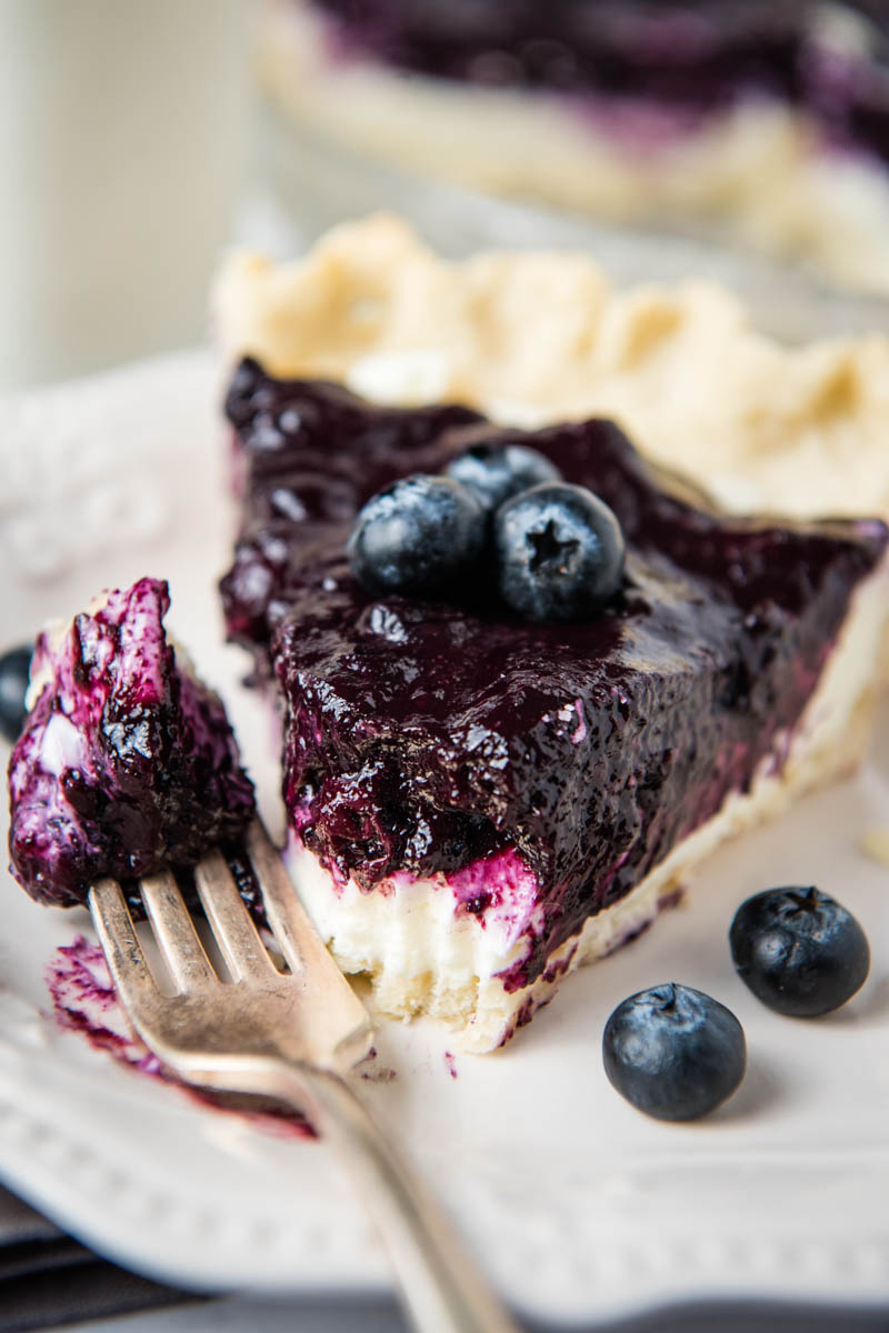 bite out of cream cheese blueberry pie slice on white plate with fork and fresh blueberries