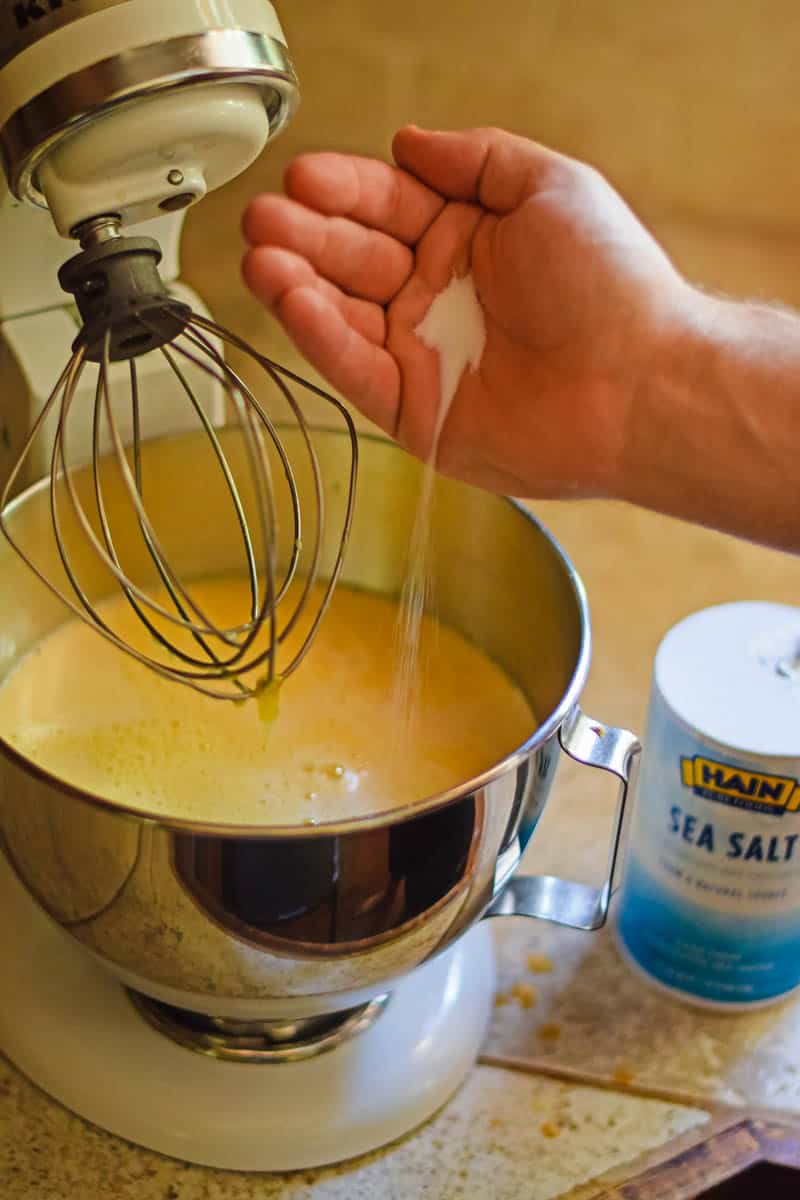 hand adding dash of salt to homemade ice cream mixture in metal bowl of KitchenAid mixer