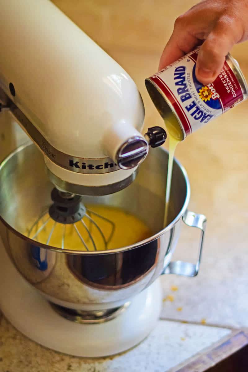 hand pouring Eagle Brand sweetened condensed milk into metal bowl of white KitchenAid mixer