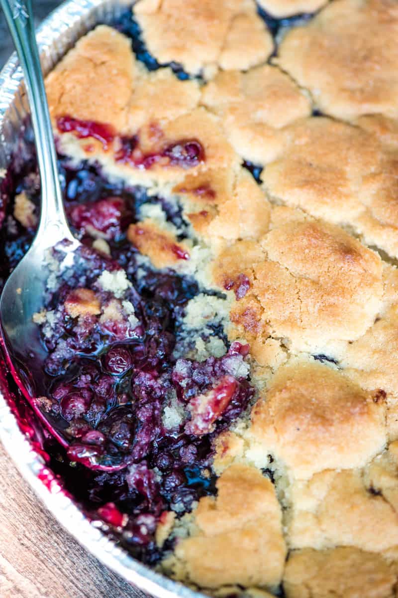 scooping cast iron blueberry cobbler out of foil pan with metal spoon