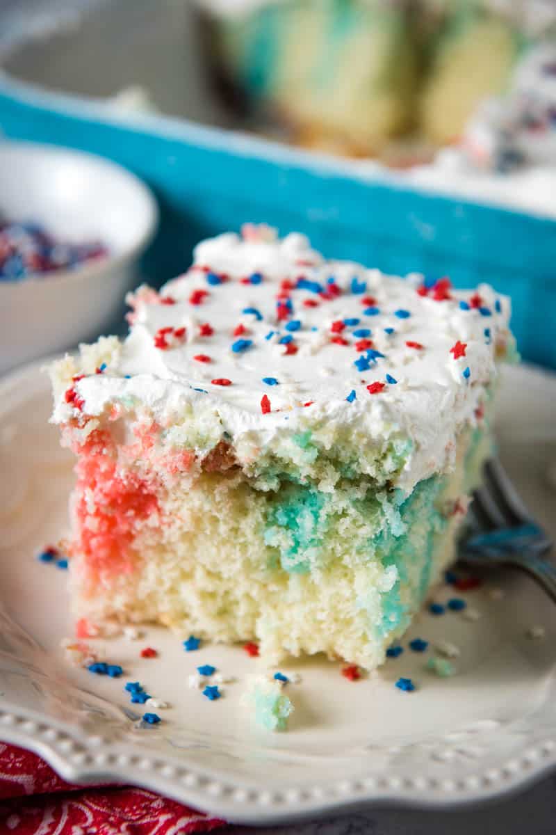 slice of red white and blue poke cake with whipped cream and sprinkles on white plate with fork