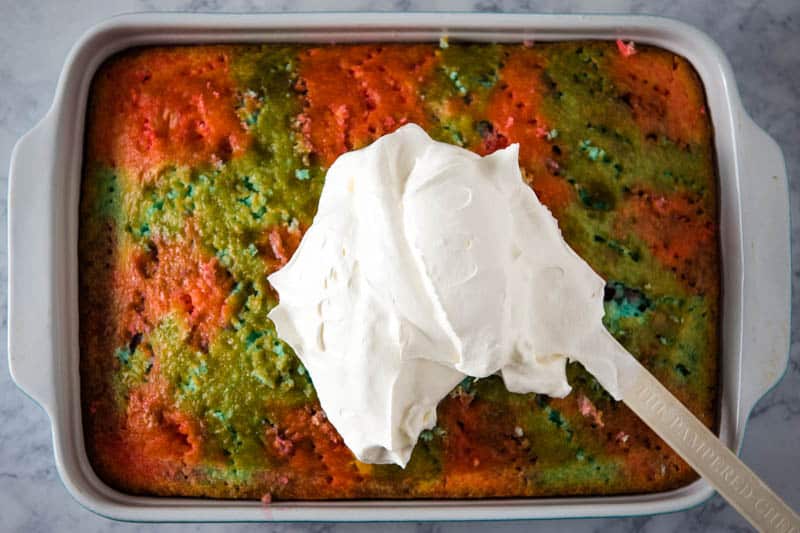 spreading Cool Whip whipped topping with spatula over top of patriotic poke cake in baking dish