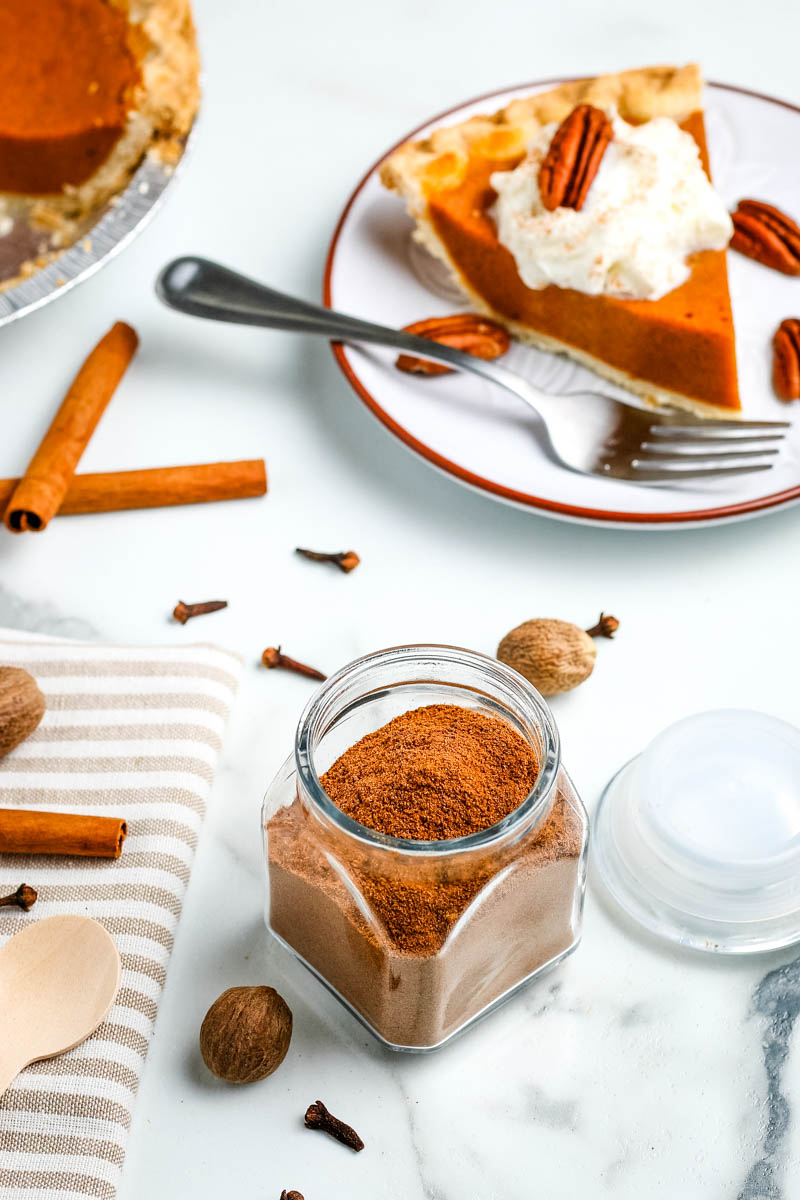 Pumpkin pie spice blend in small glass jar on white marble countertop and slice of pumpkin pie on red rimmed white plate with fork