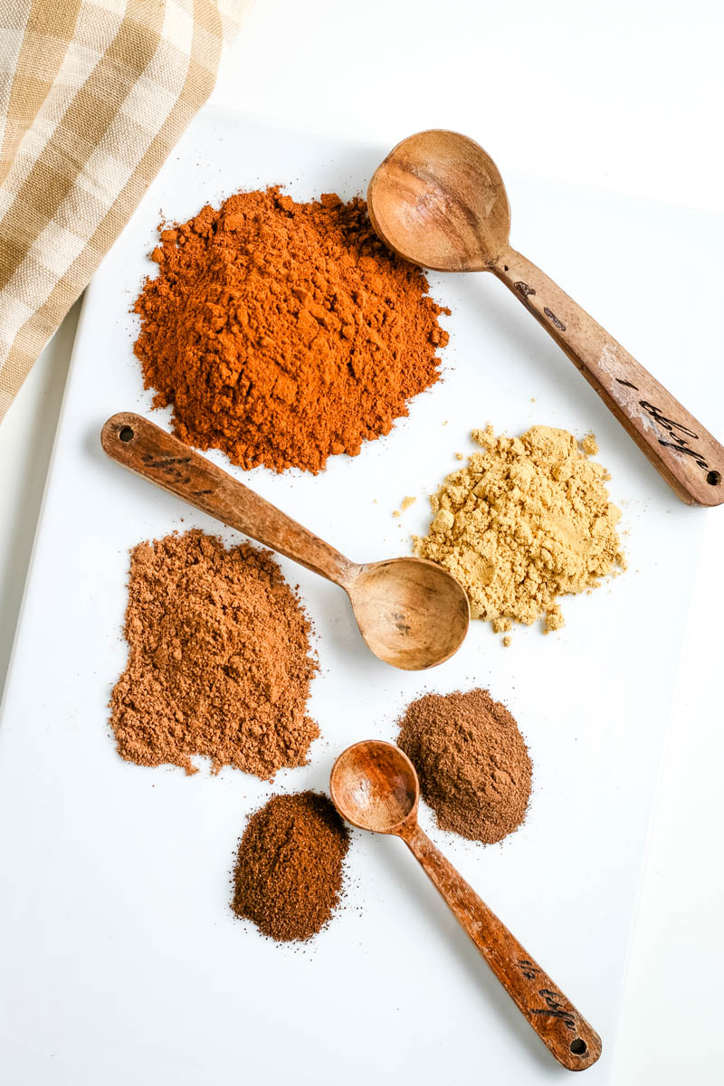 spices and seasonings used to make pumpkin spice seasoning, on white countertop with small wooden spoons and tan checkered gingham towel