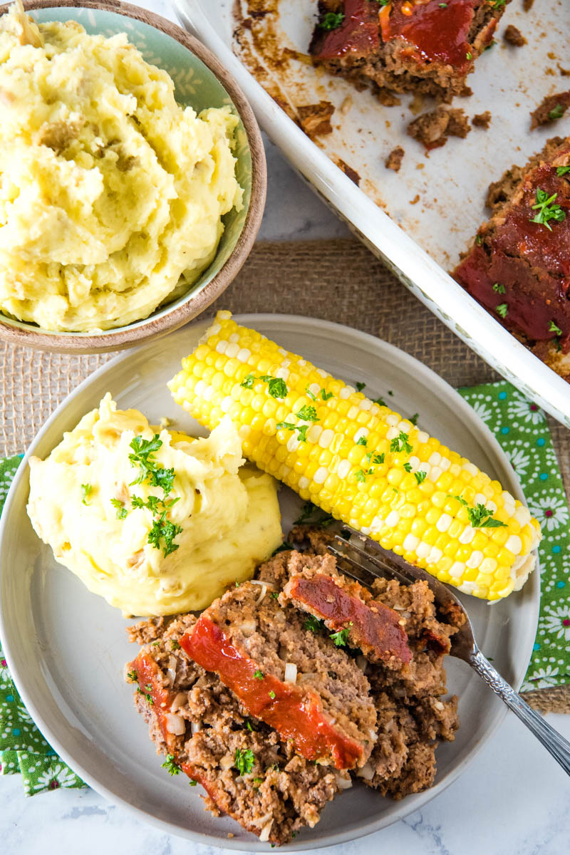 above gray plate of classic meatloaf with mashed potatoes and corn on the cob, with fork, on green and white flowered linen and tan burlap