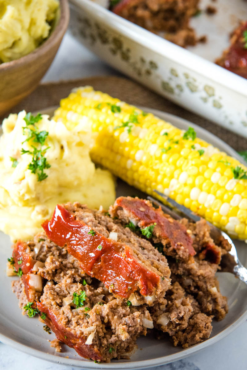 classic meatloaf sliced and served on gray plate with mashed potatoes and corn on the cob