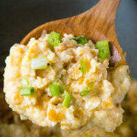 wooden spoonful of CrockPot funeral potatoes with green onions over gray slow cooker with casserole