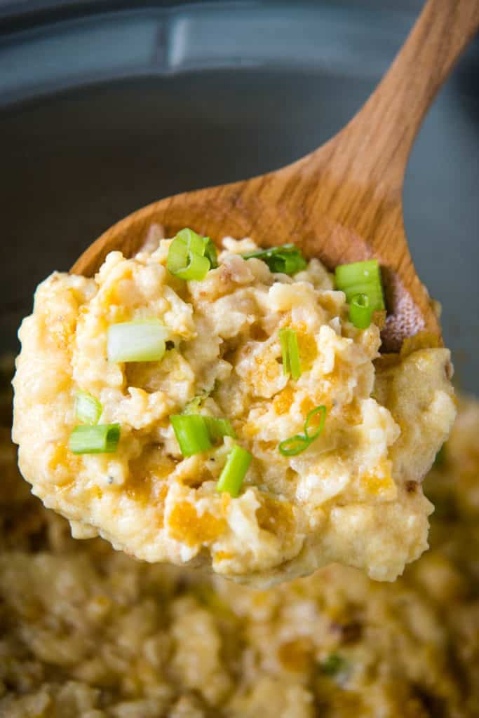 wooden spoonful of CrockPot funeral potatoes with green onions over gray slow cooker with casserole