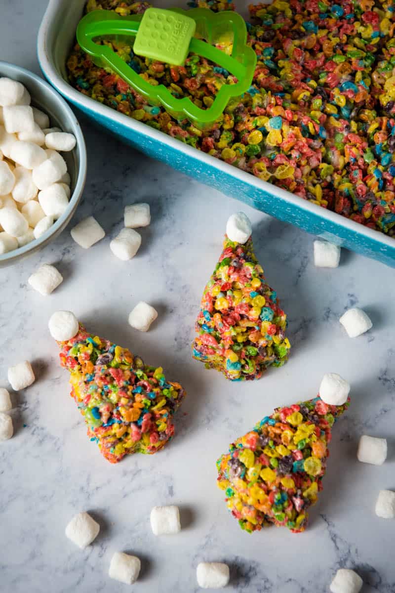 Fruity Pebble crispy treats Christmas trees laying on white marble countertop with blue pan of crispy treats and bowl of mini marshmallows