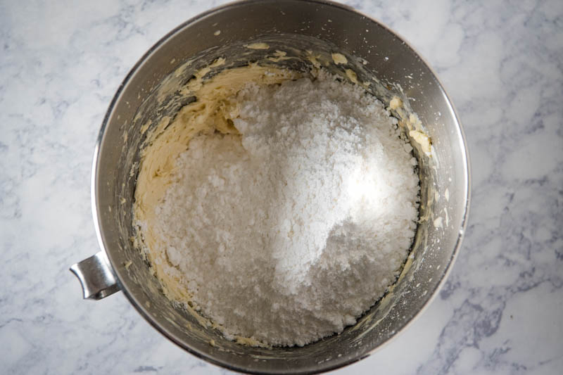 mixing powdered sugar into buttercream frosting in KitchenAid mixing bowl on white marble countertop
