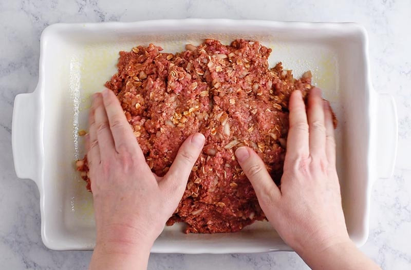 hands shaping oatmeal meatloaf in white baking dish on white marble countertop